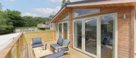 Une terrasse en bois avec des meubles d’extérieur, une porte vitrée donnant sur un patio abrité et une vue sur un paysage verdoyant luxuriant.