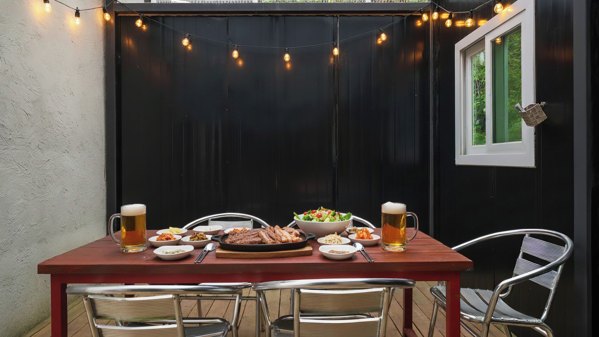 An outdoor dining area with a table set for a meal, surrounded by chairs, and string lights overhead.