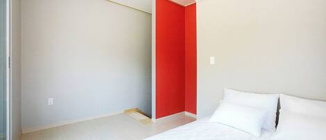 Un dormitorio con cama, almohadas blancas, una pared de acento roja y un equipo de aire acondicionado.