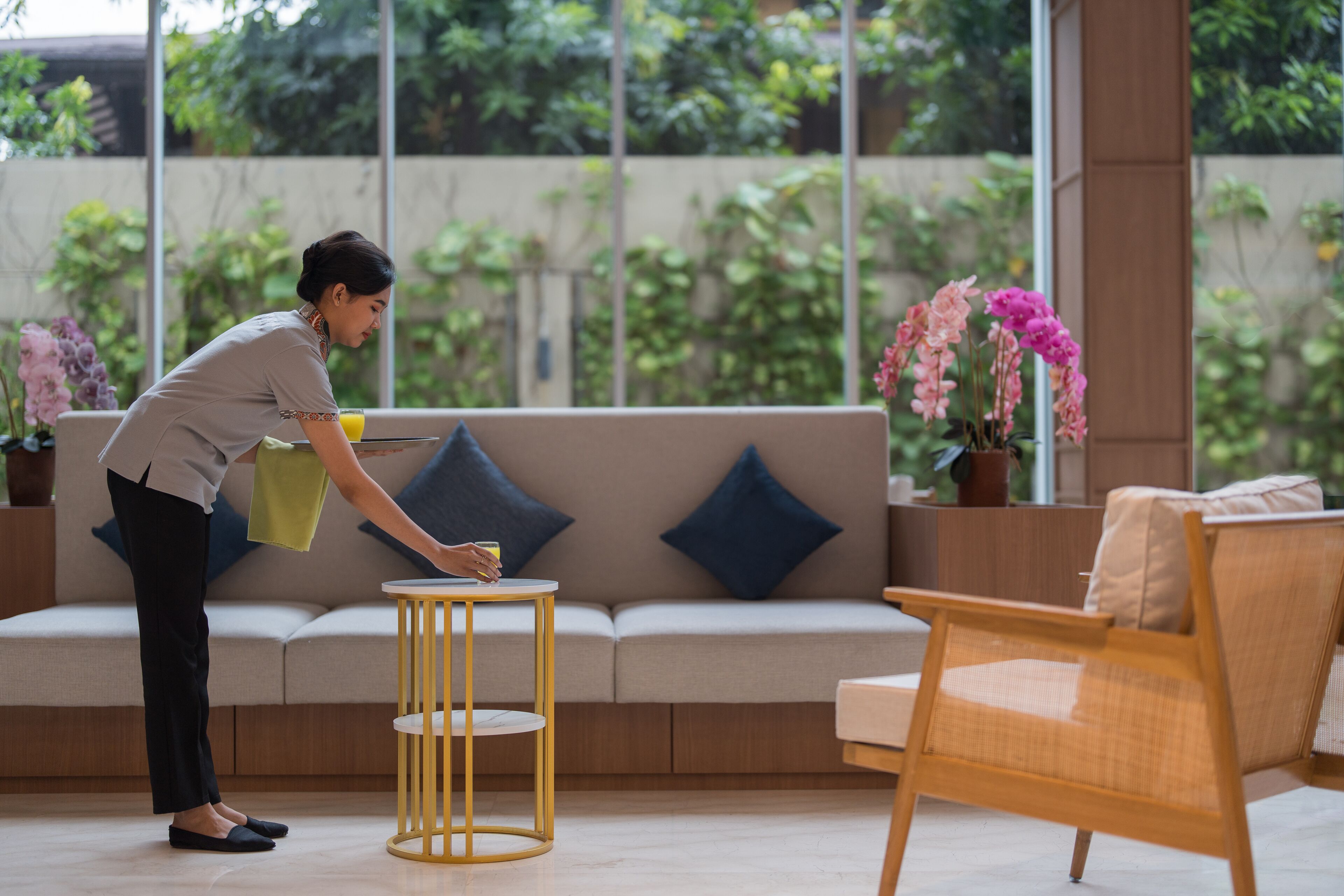 A person cleaning a round, gold-framed table in a modern living room with a beige sofa, blue cushions, and a wicker chair.