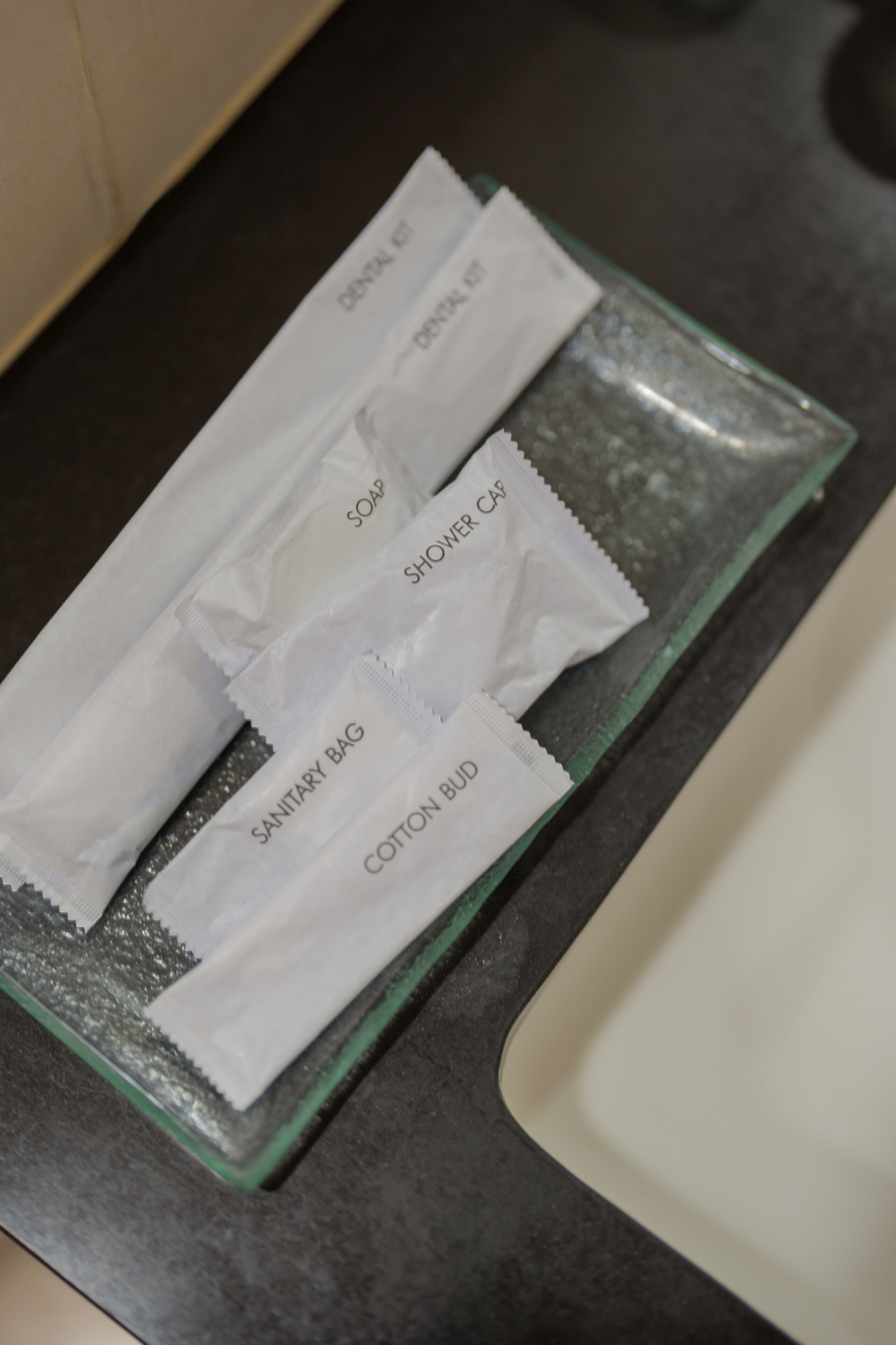 Ein Badezimmer-Waschtisch mit einer Glasablage, auf der verschiedene Pflegeprodukte liegen, darunter eine Hygiene-Tasche, Wattestäbchen, Seife und Duschcreme.