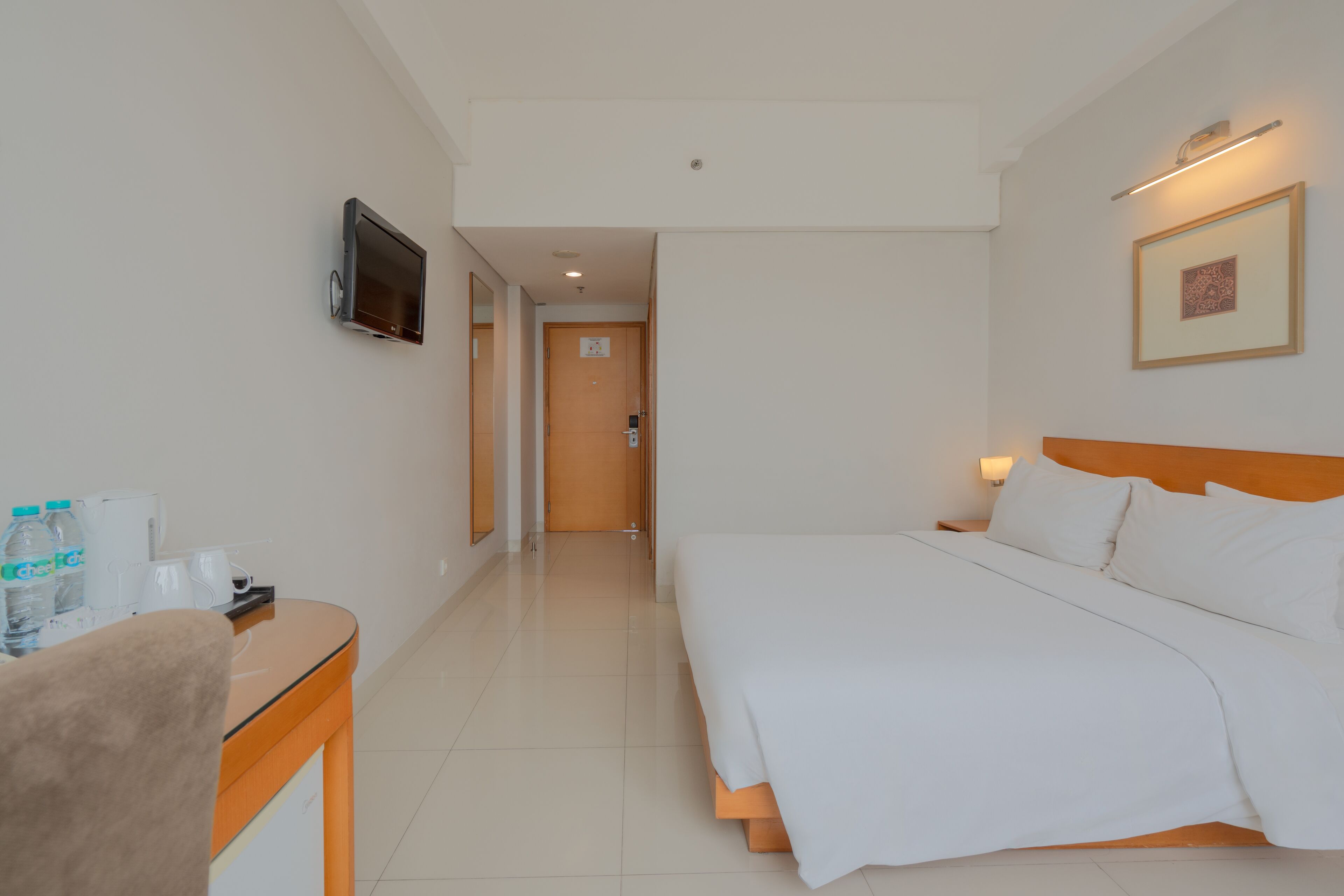 A hotel room with a bed, a desk with toiletries, a TV mounted on the wall, and a framed picture above the bed.