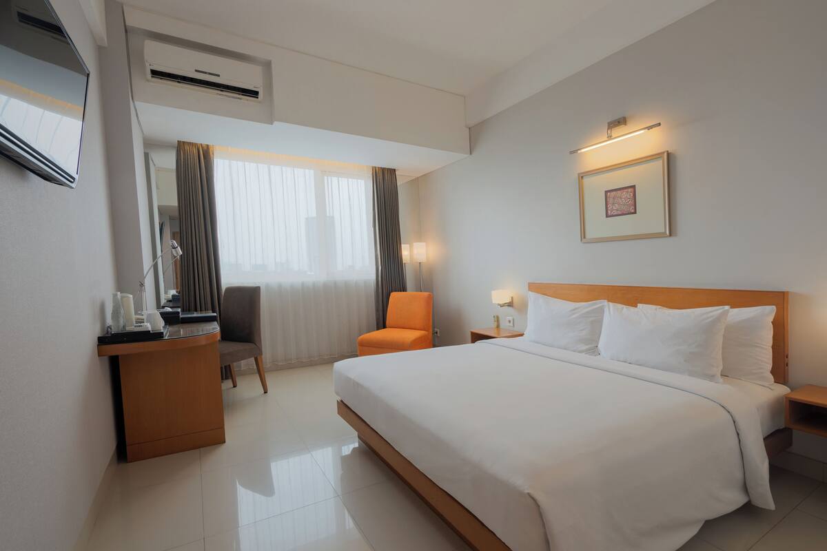 Habitación de hotel con una cama grande, un escritorio con silla, una mesita de noche con lámpara y una ventana con cortinas.