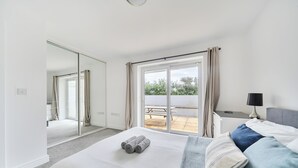 A modern bedroom with a large bed, a bedside table with a lamp, a mirror, and a sliding glass door leading to an outdoor patio. - Spacious 3 Bed House - Sleeps 6 - Parking - Wifi (Newquay)