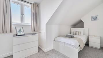 Um quarto com uma cama, mesas de cabeceira, cômoda, janela com cortinas e um quadro emoldurado sobre a cômoda.