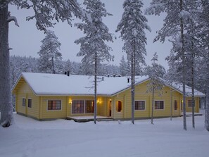 A yellow cabin with a snow-covered roof, surrounded by tall, snow-laden trees.