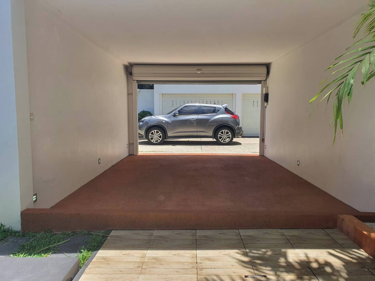 Un garage moderne avec une voiture garée à l’intérieur, une rampe menant à la porte du garage et un pot de fleurs placé sur la droite.