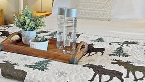 Una cama bien tendida con colcha blanca, cabecera de madera y mesita de noche con lámpara y bandeja con planta y dos botellas de agua.