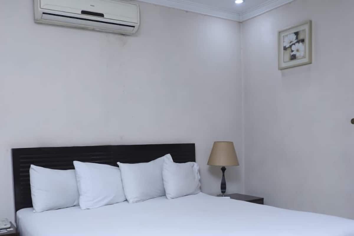 A large bed with white bedding, a dark headboard, a bedside lamp, and an air conditioning unit on the wall.