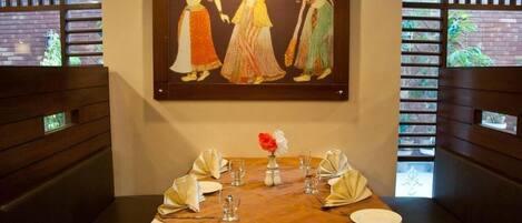 A dining area with a table set for four, a framed painting above, and a view through a window with a brick wall.