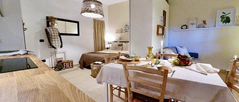 A compact, well-lit room with a dining table set for two, a bed with a headboard, and a small kitchenette area.