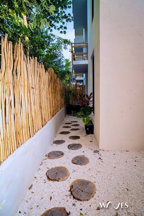 Un sendero estrecho con aberturas circulares en el suelo, bordeado por una cerca de bambú y plantas en macetas.