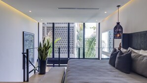 A modern bedroom with a large bed, a bench, a potted plant, and a view of tall buildings through large windows.