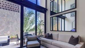 A modern living room with a beige sofa, a small black ottoman, and a glass door leading to an outdoor area with a stone wall and plants.