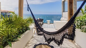 A hammock on a balcony overlooking a pool and ocean, with a white sofa and a small table nearby.