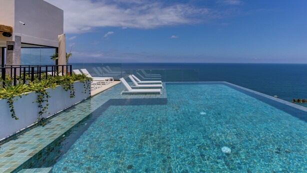 A clear blue infinity pool with white lounge chairs, a white building with a glass facade, and a view of the ocean.