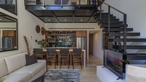 A modern kitchen with a central island, a dining area with wooden chairs, a sofa, and a staircase with a metal railing.