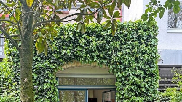 Un espace extérieur accueillant, aménagé avec des sièges confortables et doté d’une porte vitrée à l’entrée, le tout entouré d’une végétation luxuriante et de plantes en pot.