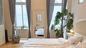 Une chambre avec un grand lit, deux fenêtres avec des rideaux gris, une plante en pot et un radiateur.