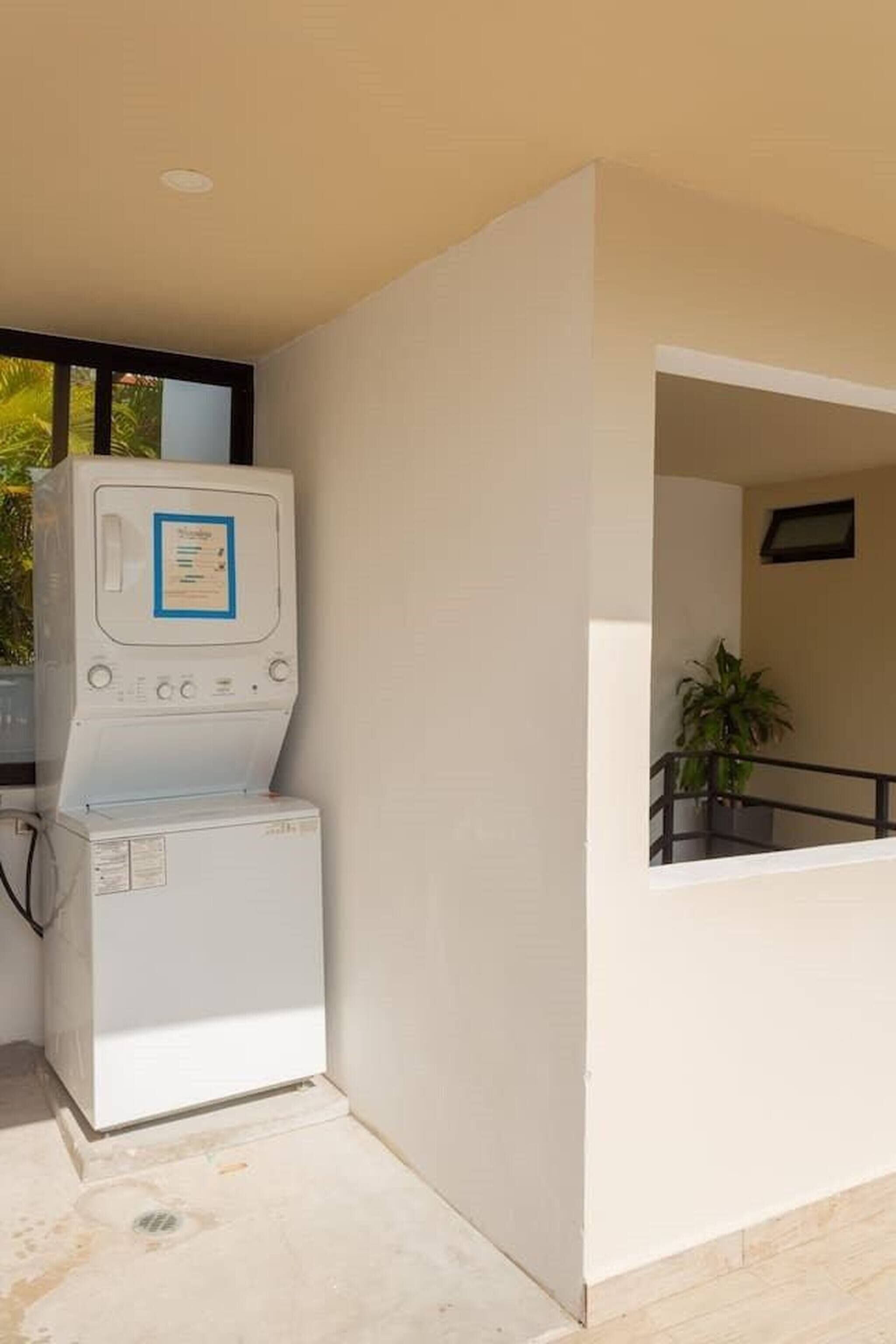 A laundry area with a washing machine and dryer, a window with a view of greenery, and a potted plant on a balcony.