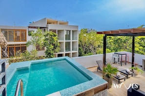 A rooftop swimming pool with a pergola and seating area, surrounded by greenery and modern buildings.
