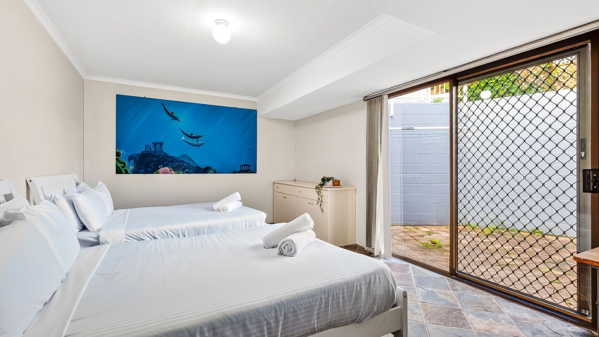 A hotel room with two beds, a large window with a view of a courtyard, and a painting of an underwater scene on the wall.