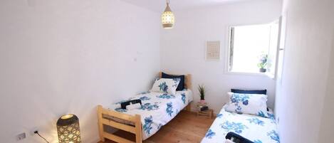 A small, well-lit room with two wooden beds, a floor lamp, and a window providing natural light.