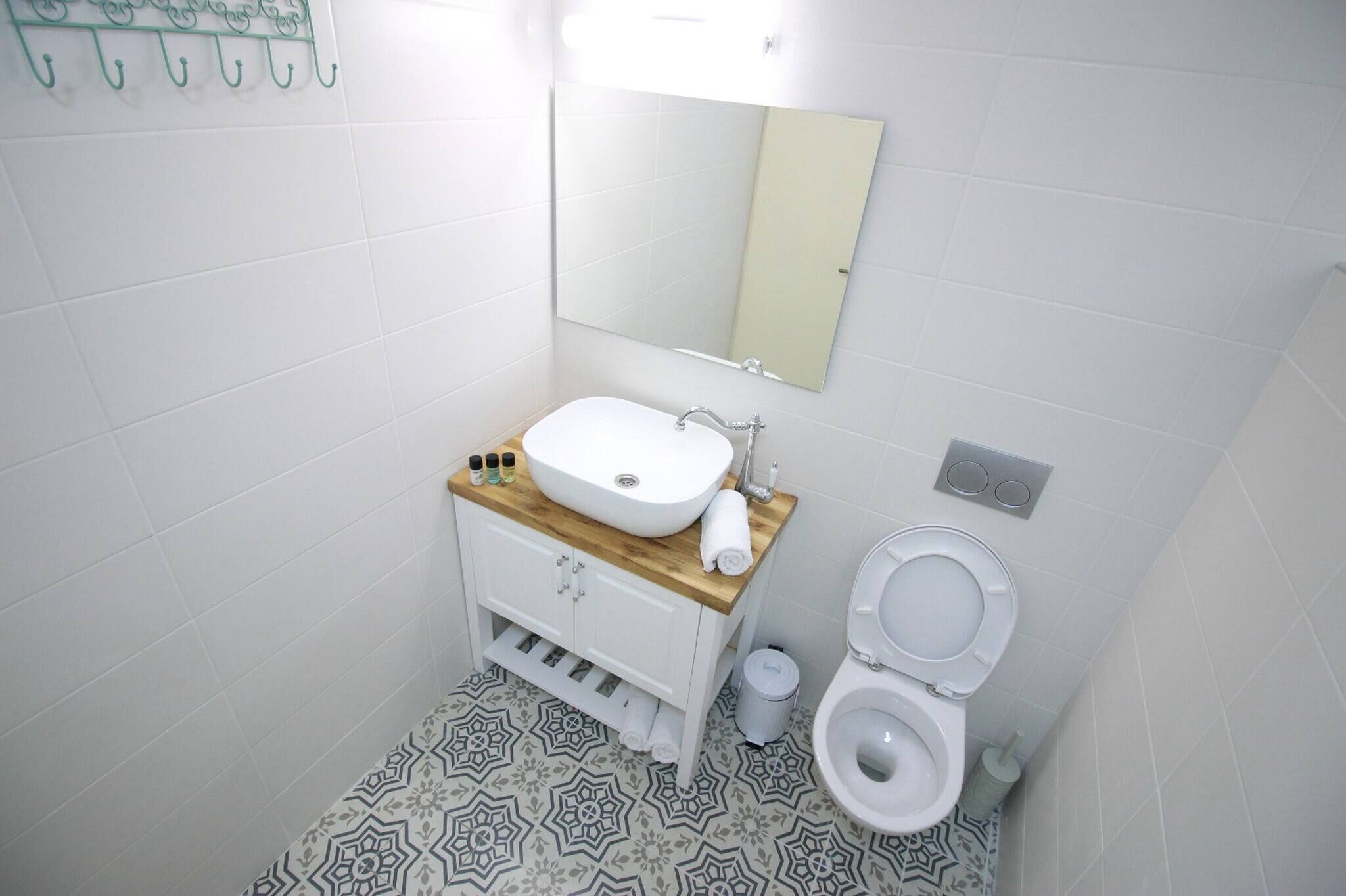 A bathroom with a white sink, wooden vanity, mirror, toilet, patterned floor, and a towel rack.