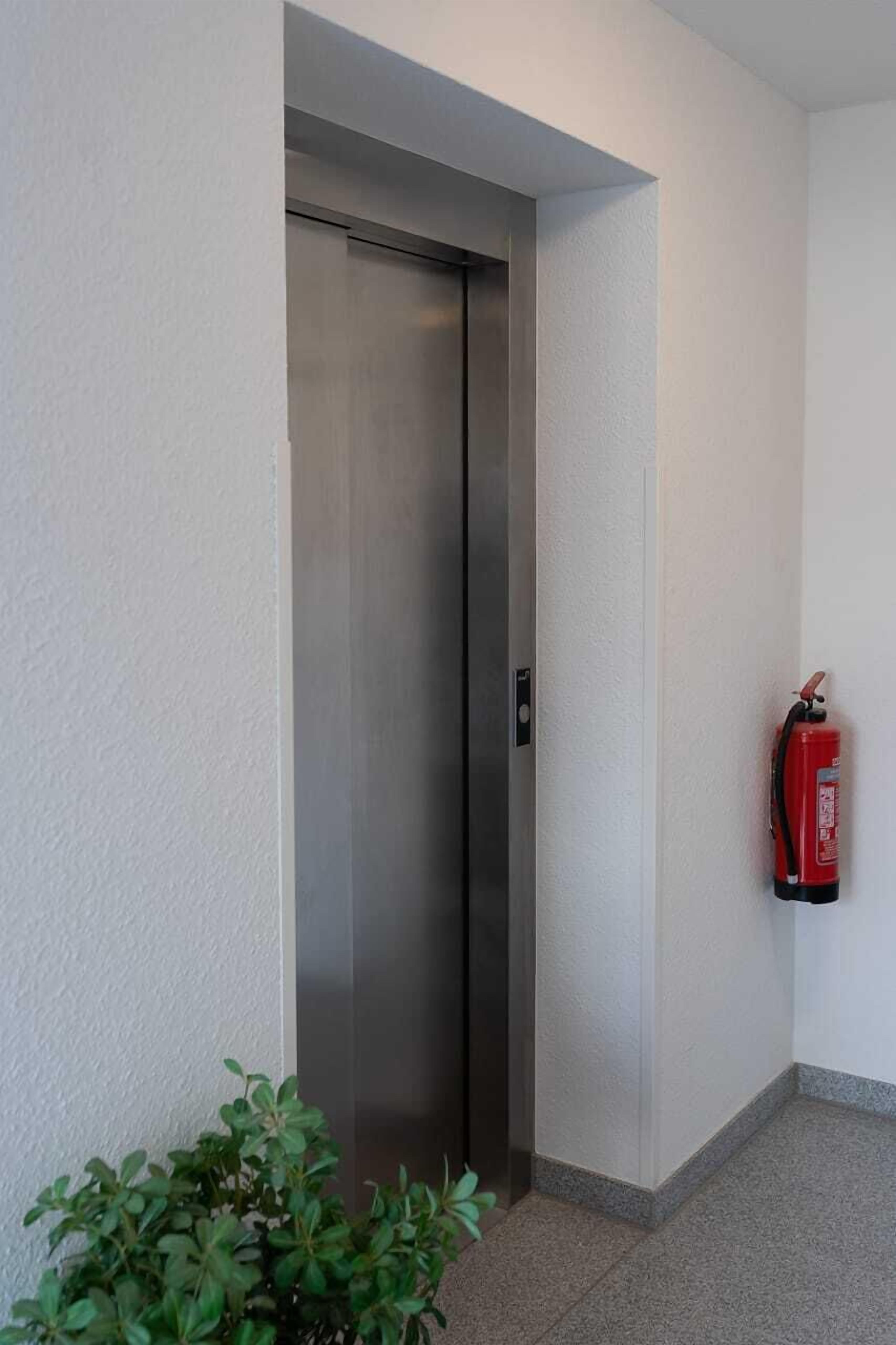 A narrow hallway with a metallic elevator and a red fire extinguisher mounted on the wall.