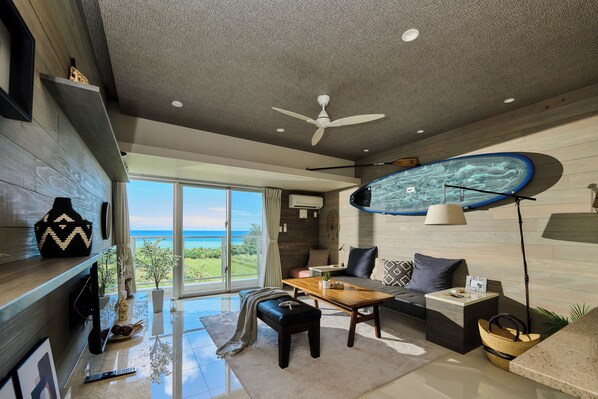 A modern living room with a large window, a ceiling fan, and a view of the ocean. - Grand blue 303 (Onna)