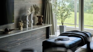 Una sala moderna con un televisor de pantalla plana, una pared de madera, un estante con objetos decorativos, un ventanal con vistas a la vegetación y un sofá de cuero.