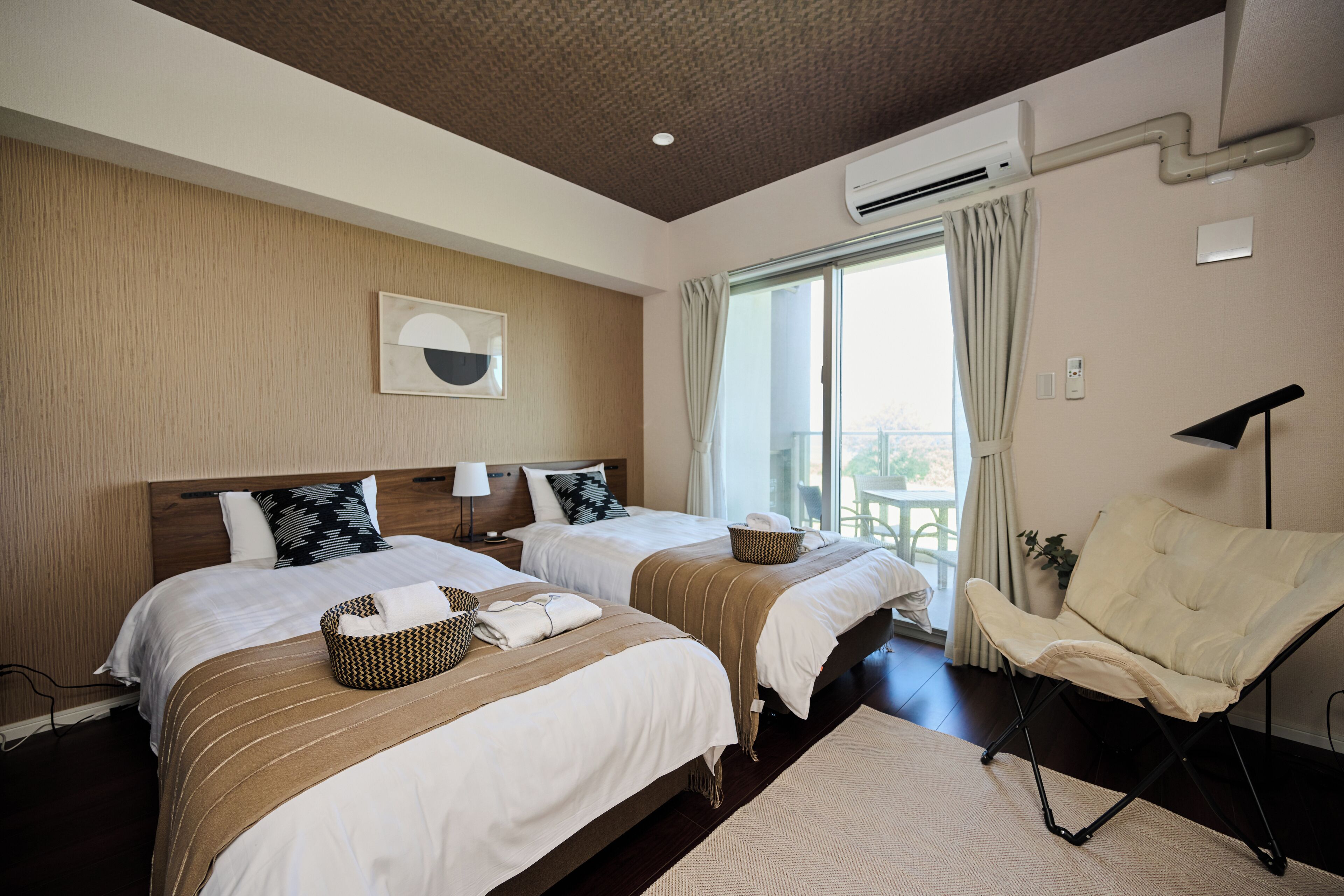A hotel room with two beds, a chair, a lamp, and a view of the outdoors.