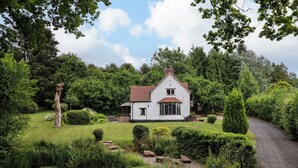 Une maison blanche avec un toit rouge, entourée d’une végétation luxuriante et d’un étang.