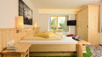A wooden bed with pillows, a bedside table with a phone, a TV on the wall, and a window with a view of greenery.