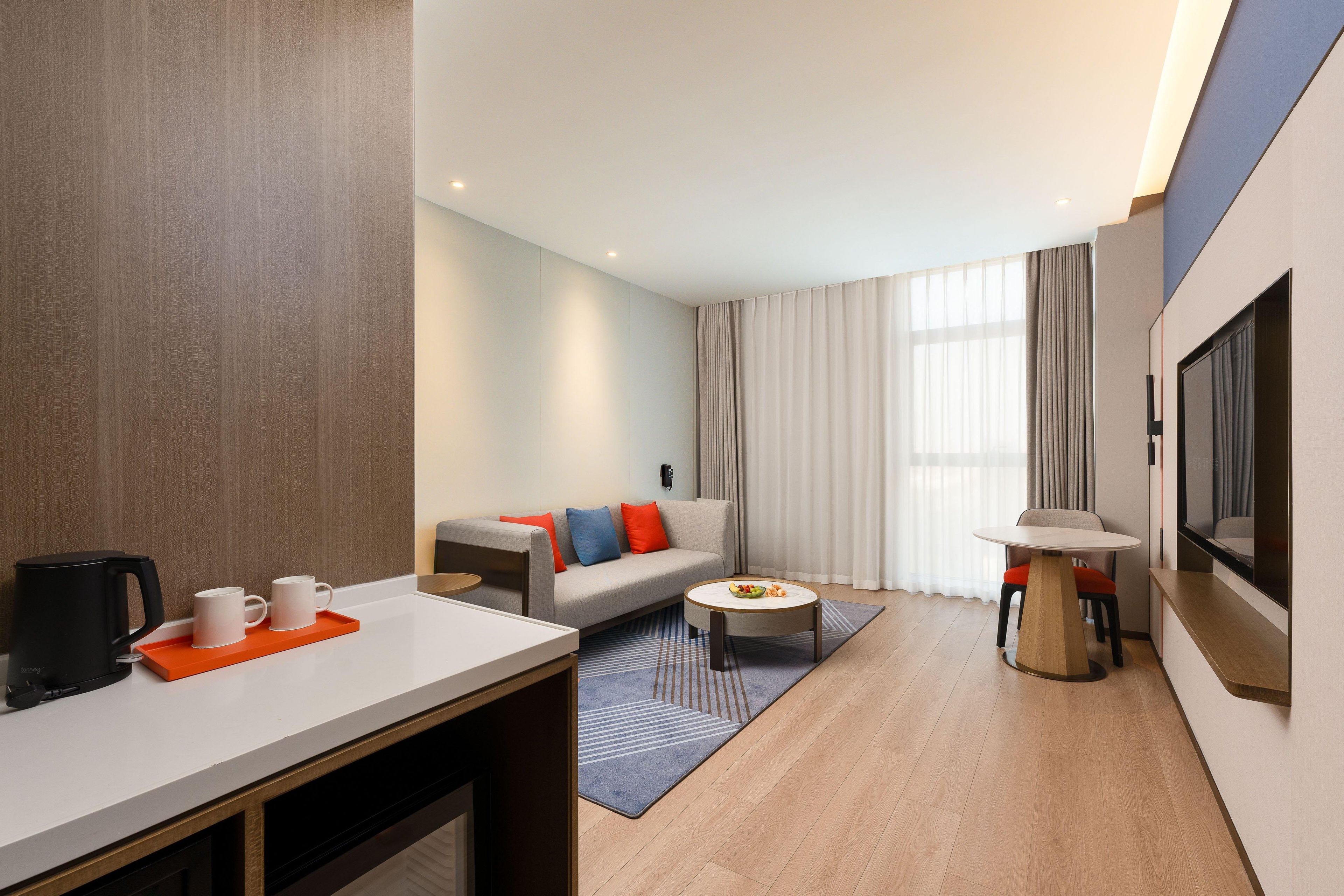 A modern hotel room with a sofa, coffee table, TV, and a dining area with a coffee maker and mugs.