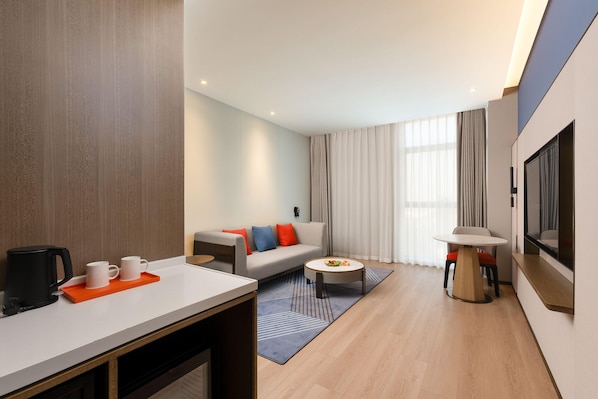 A modern hotel room with a sofa, coffee table, TV, and a dining area with a coffee maker and mugs.