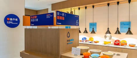 Zona de recepción de un hotel con un mostrador que exhibe varios recipientes con comida, un cartel para desayuno exprés y un cartel en la pared con opciones de bebidas.