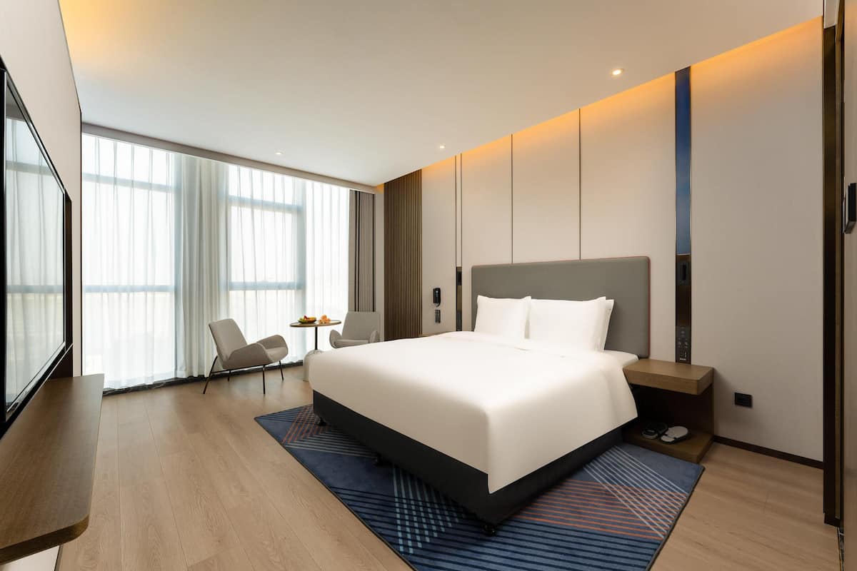 A modern hotel room with a large bed, a desk, a chair, and a view of the city through large windows.