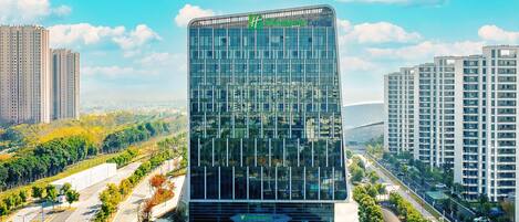 Un edificio moderno de vidrio, con un logotipo verde, rodeado de estacionamiento y vegetación.