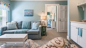 A cozy living room with a grey sofa, patterned ottoman, and a small white table. A painting hangs on the wall, and a lamp is on a side table. - Beautiful inn, Lighthouse Landing. #3 Sea Glass (Ogunquit)
