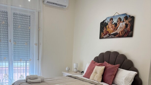 A bedroom with a large bed, a nightstand, a lamp, a painting, and an air conditioning unit. - Apartment bari center (Bari)