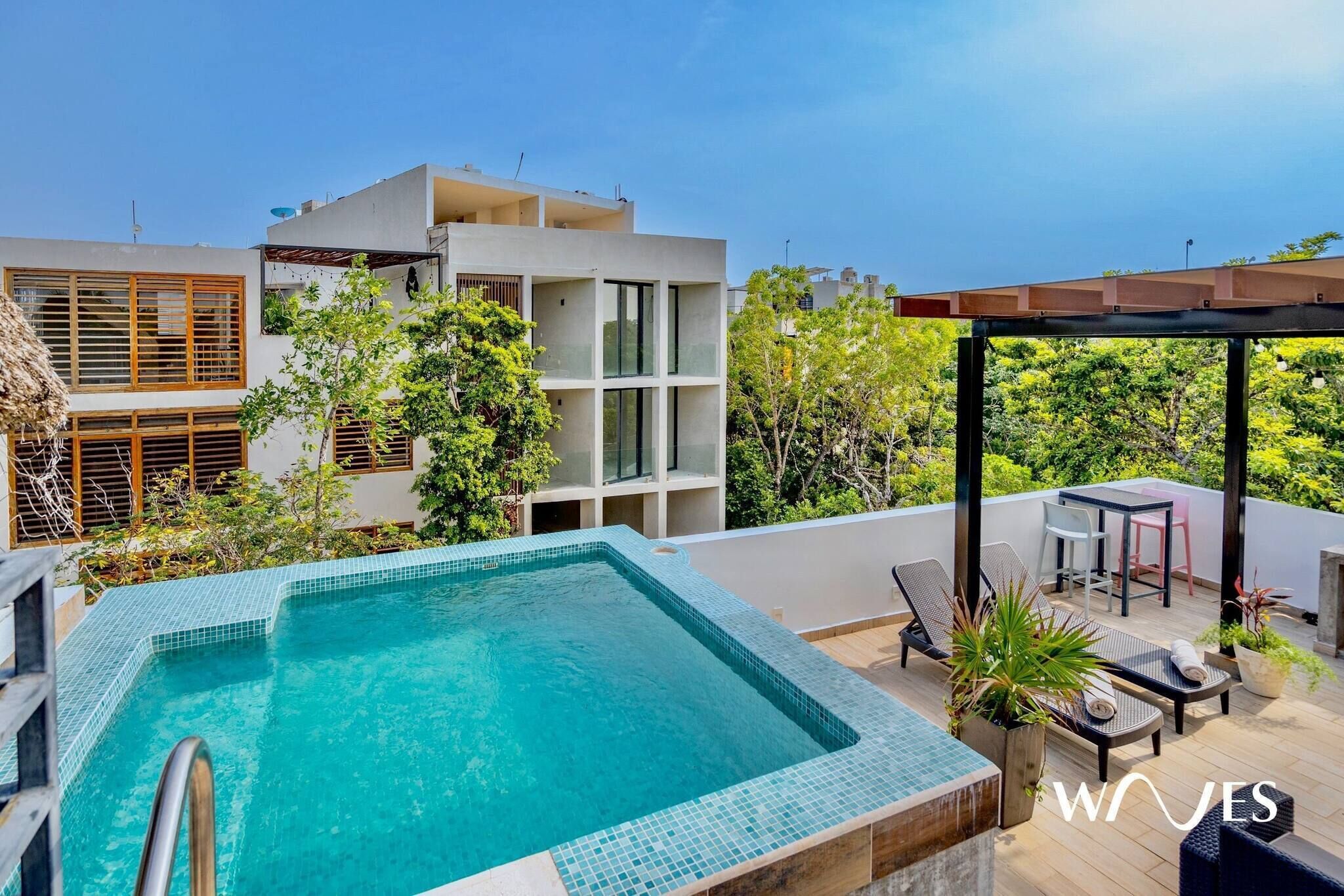 A rooftop swimming pool with a pergola and seating area, surrounded by greenery and modern buildings.