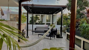 A covered outdoor seating area with a hammock, a sofa, and a table, surrounded by potted plants and a view of a building. - Room in Apartment - Spacious Great Located Amazing Terrace (Tulum)