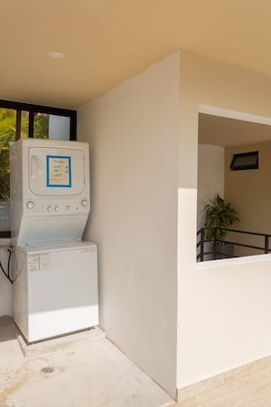 Zona de lavandería con lavadora y secadora, ventana con vista a vegetación y planta en maceta en un balcón.