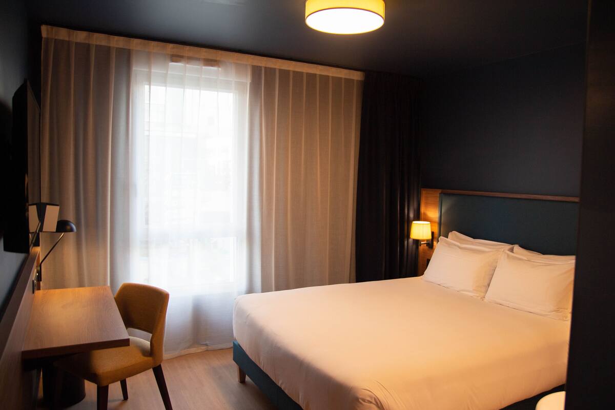 Habitación de hotel con cama, escritorio, silla, lámpara y ventana con cortinas.