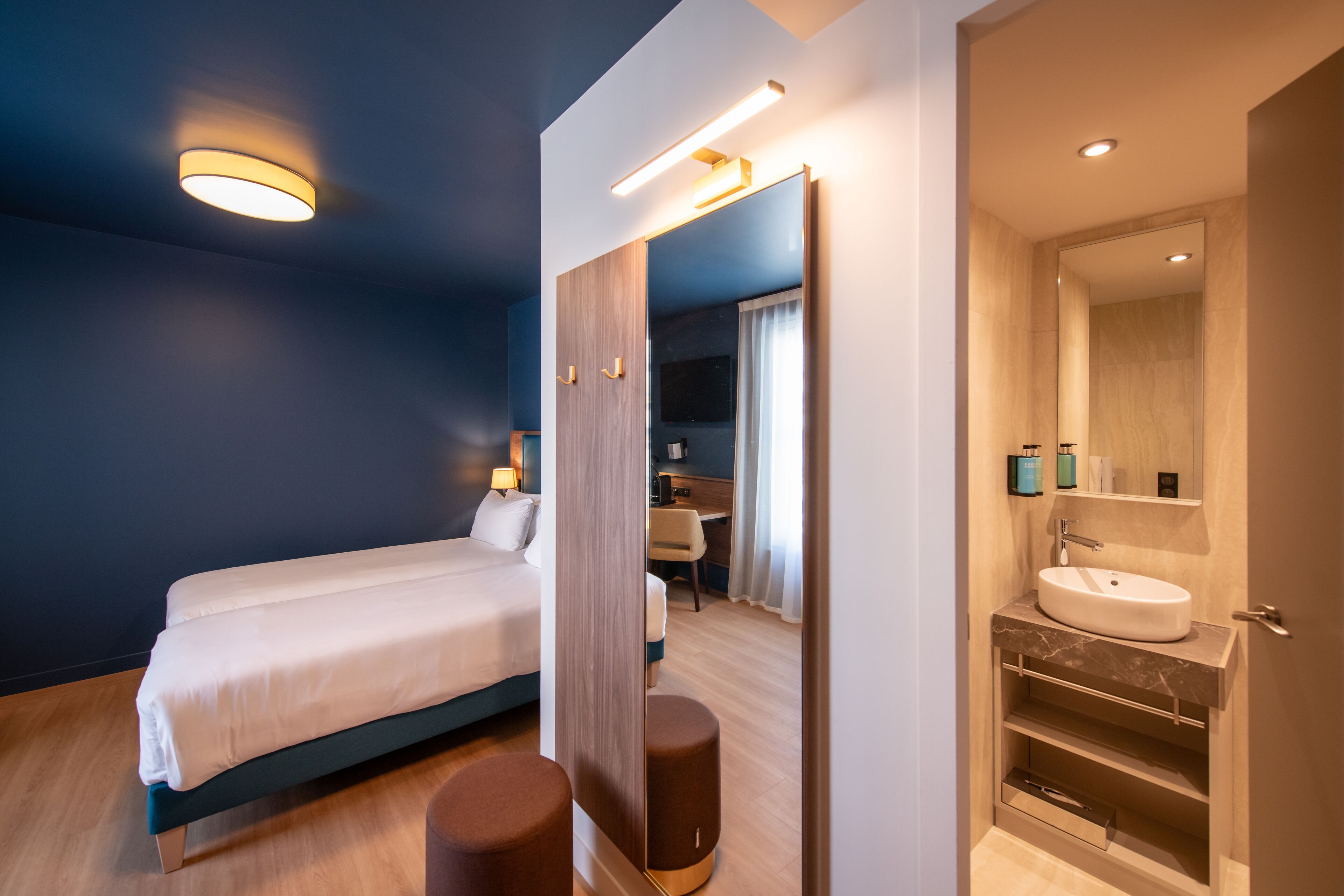 A modern hotel room with a bed, a sitting area, a bathroom with a sink and mirror, and a desk with a chair.