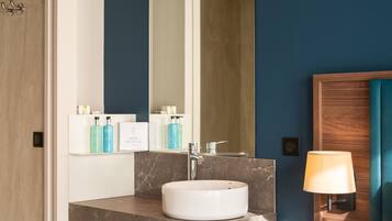 A modern bathroom with a white sink, marble countertop, and a large mirror. There are shelves with toiletries and a wooden nightstand with a lamp.