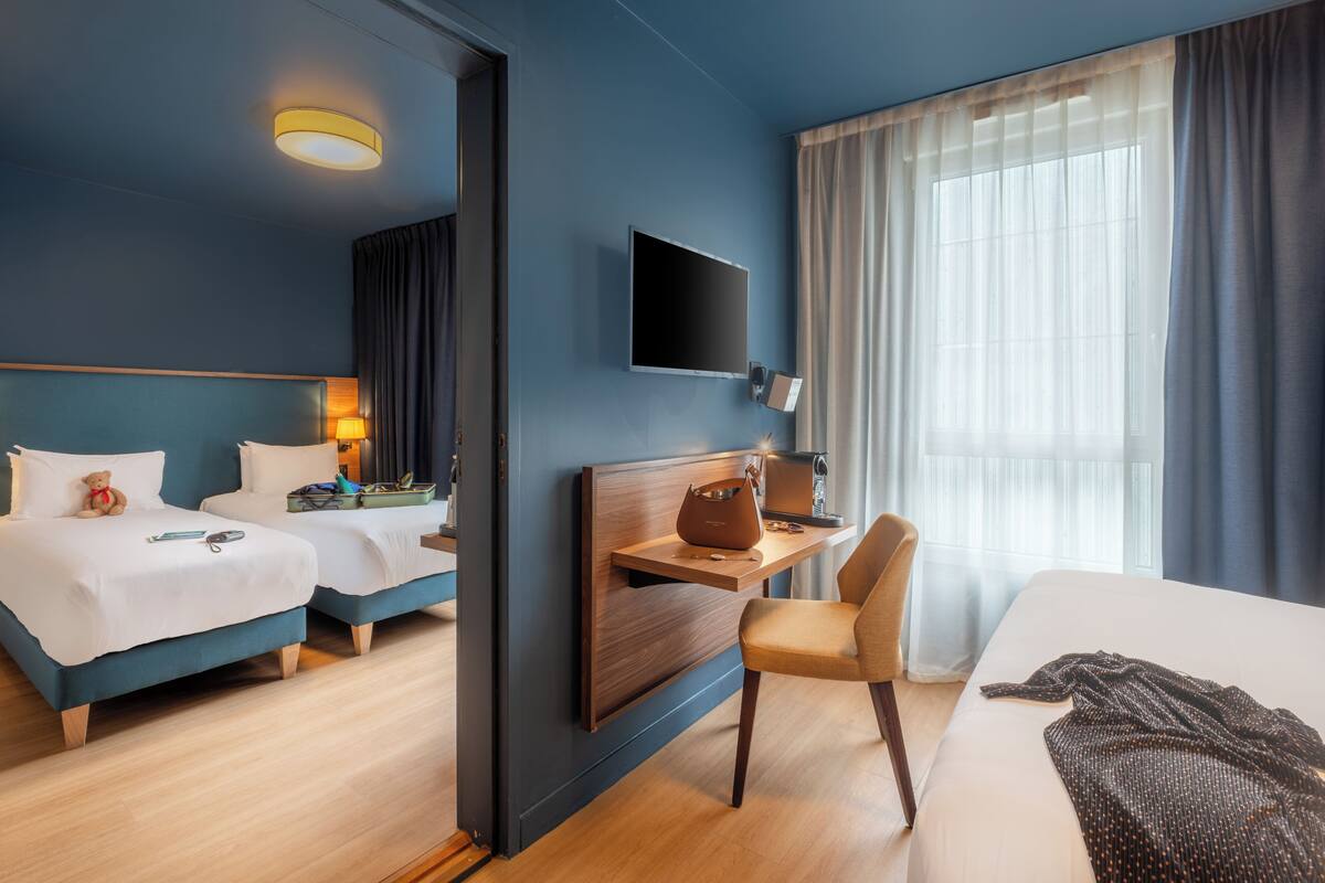 Habitación de hotel con dos camas, un escritorio, una silla, un televisor y un ventanal con cortinas.
