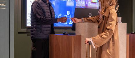 A reception area with a digital screen displaying hotel information, a wooden reception desk, and a green suitcase.
