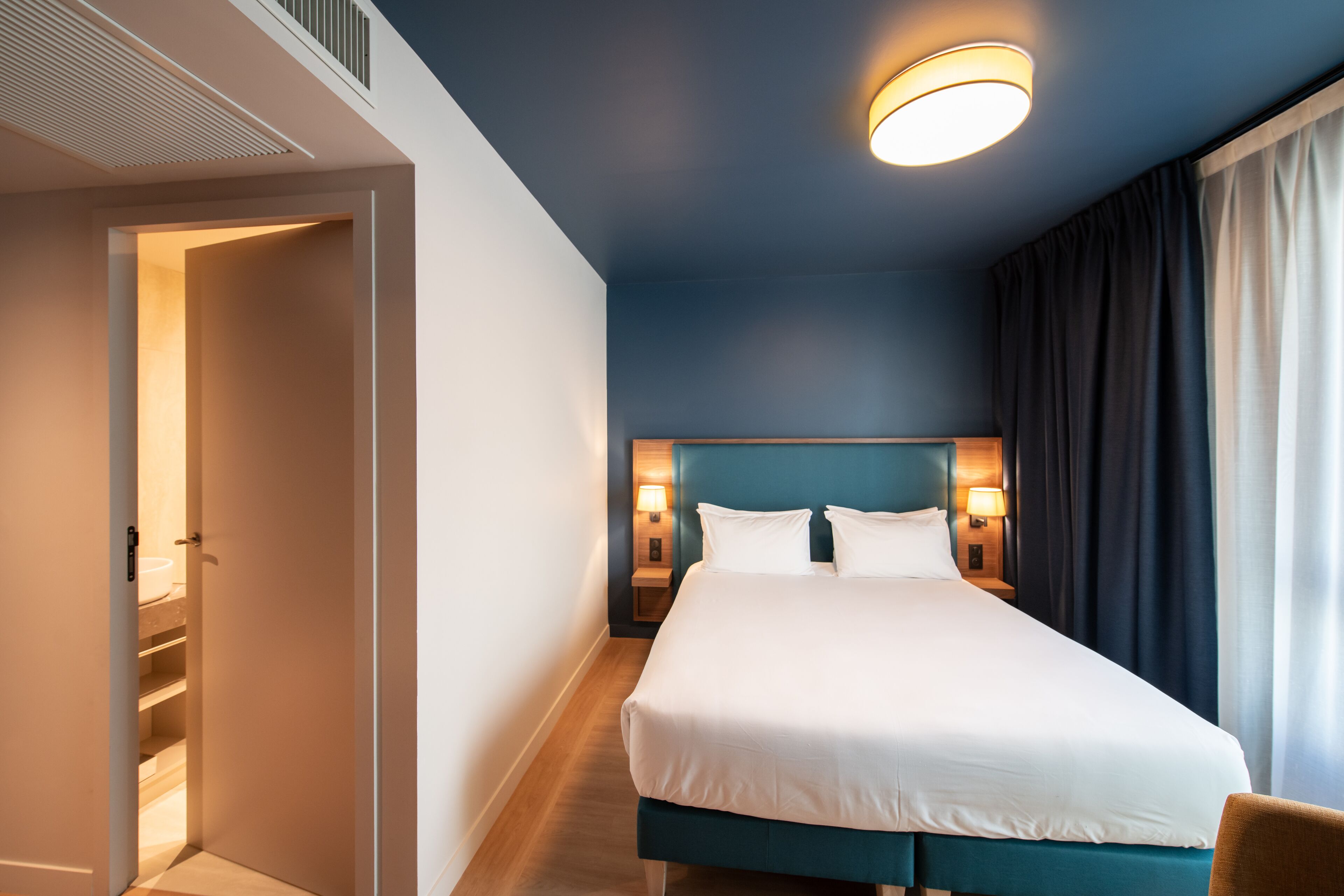 Habitación de hotel con una cama grande, dos lámparas de noche, una lámpara de techo y vistas a una ventana con cortinas.